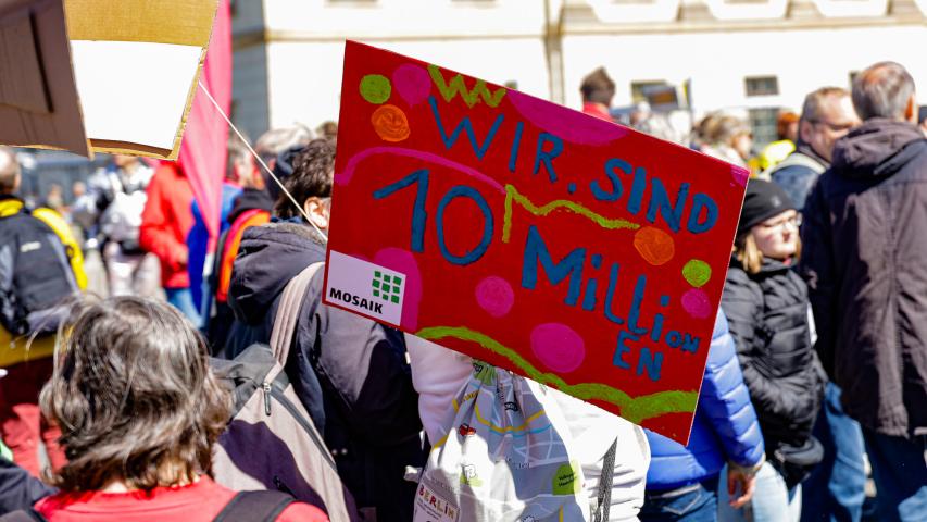 Ein Protestschild "Wir sind 10 Millionen" und das Mosaik-Logo